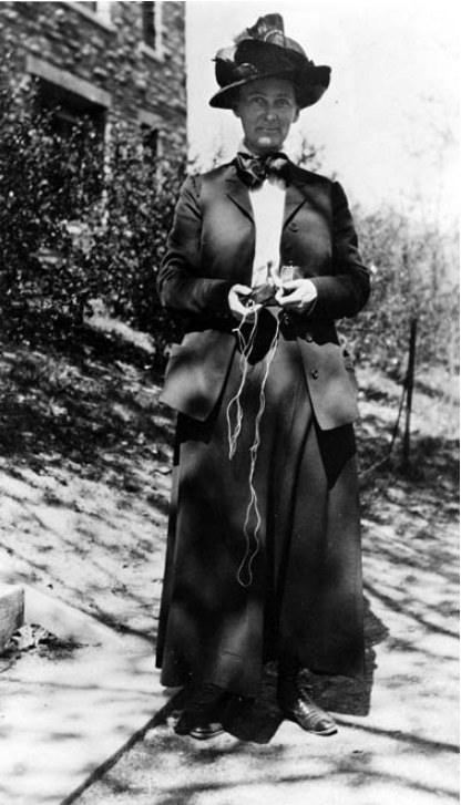 Florence Bascom wearing a jacket and a bow around her neck. She also has a large hat. She is holding a compass in her hands.