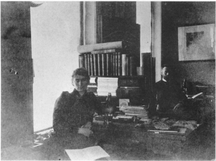 A young woman and a bearded man sit at a lab desk with books and microscopes
