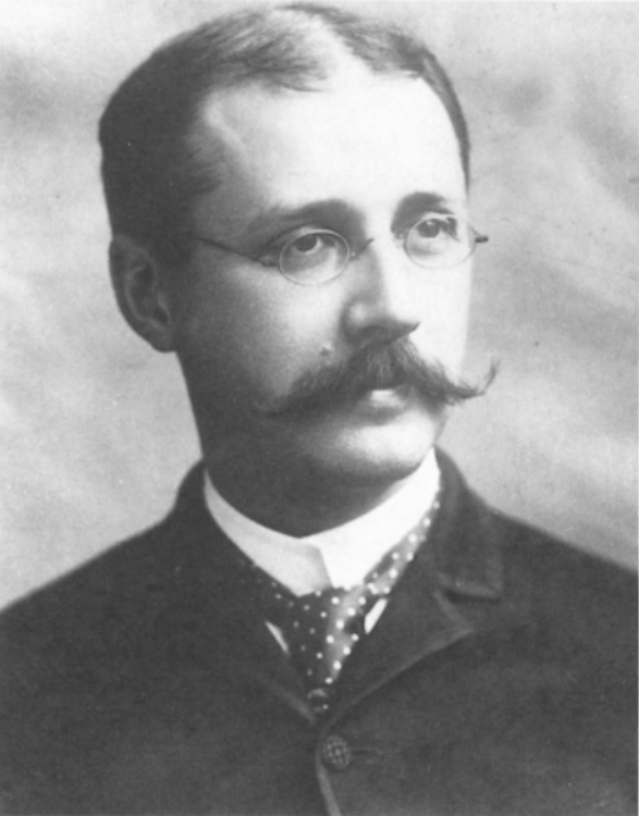 A photograph of a man wearing glasses and a long, curled mustache. Wearing old style formal clothes
