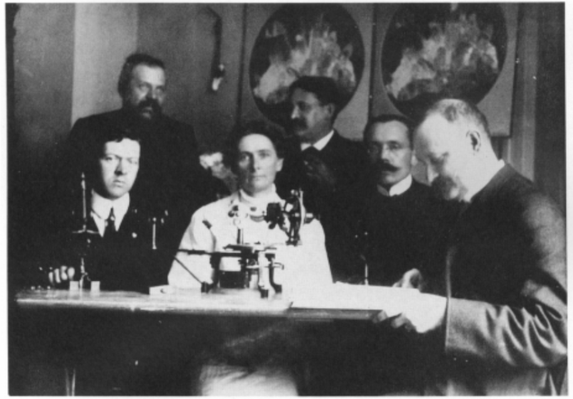 Photograph of five men and one woman wearing old style clothes sitting in front of a scientific instrument.