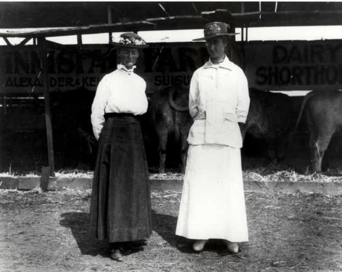 Two women in older style dresses and hats with flowers stand in front of cattle at a farm show