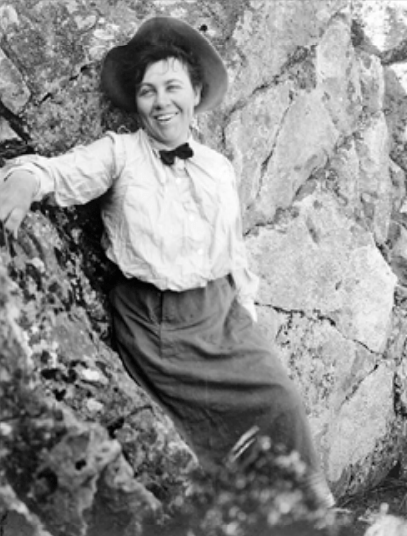 A smiling woman leaning up against a rockface wearing a hat, blouse, skirt, and small dark bowtie