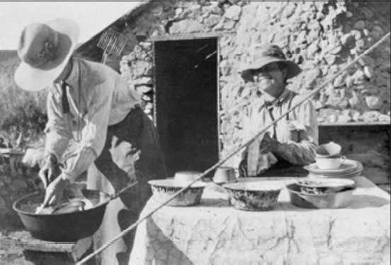 Two women with hats cleaning dishes at a campsite with a cabin behind them