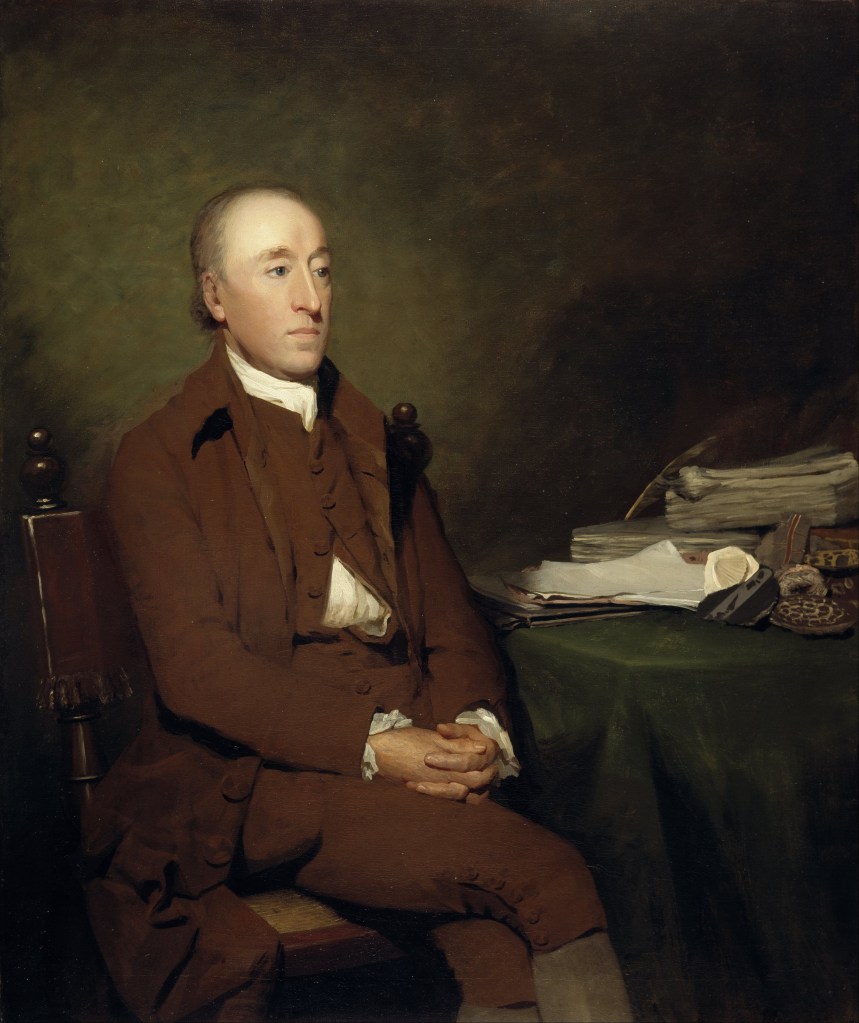 James Hutton sitting at a desk. He has short hair, and is wearing a brown coat and vest with hands folded on his lap.